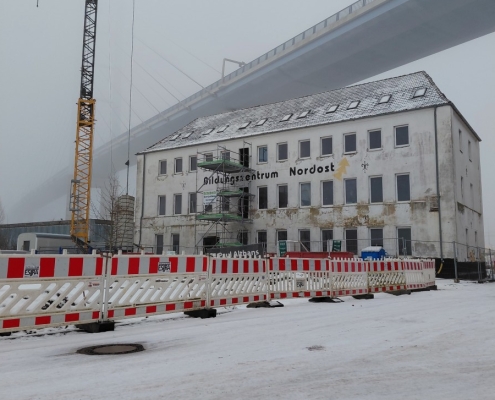 Die neue Betriebsstätte der Stralsunder Werkstätten mitten im Baugeschehen befindet sich im dichten Nebel. Im Vordergrund sind Bauzäune, ein Kran sowie ein Baugerüst zu sehen.