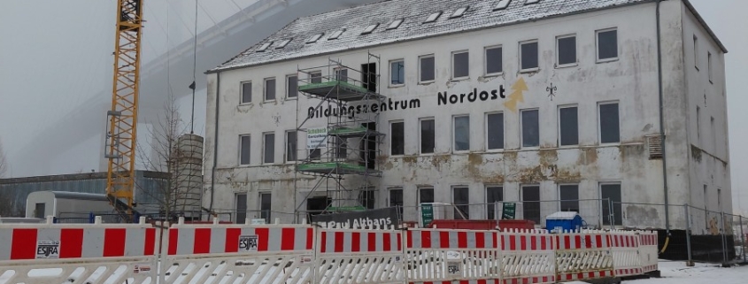 Die neue Betriebsstätte der Stralsunder Werkstätten mitten im Baugeschehen befindet sich im dichten Nebel. Im Vordergrund sind Bauzäune, ein Kran sowie ein Baugerüst zu sehen.