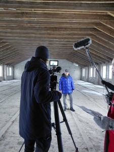 Im Vordergrund befindet sich ein Kameramann an seiner Kamera. Im Hintergrund steht eine Person vor der Kamera inmitten der im Bau befindlichen zukünftigen Betriebsstätte der Stralsunder Werkstätten.