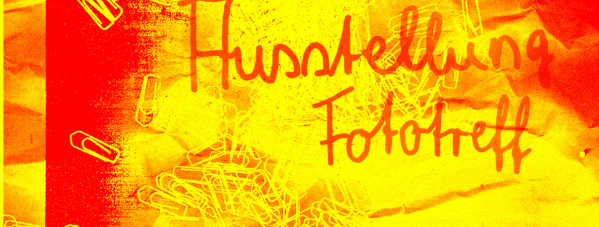 Ein Plakat in rot, orange und gelb mit der Aufschrift "Ausstellung Fototreff". Im Hintergrund sind Büroklammern zu sehen.